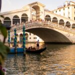 A gondola navigates the Grand Canal with Rialto Bridge lit by the setting sun in Venice, Italy.